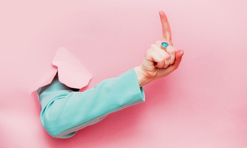 Eine Hand in einem hellblauen Ärmel ragt durch einen zerrissenen, rosa Hintergrund. Der Zeigefinger ist nach oben gestreckt, und die Hand trägt einen auffälligen Ring mit einem großen, blauen Stein