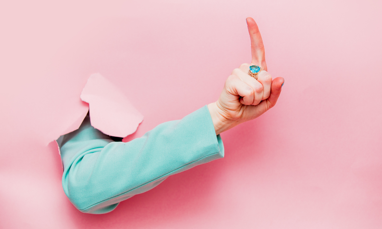 Eine Hand in einem hellblauen Ärmel ragt durch einen zerrissenen, rosa Hintergrund. Der Zeigefinger ist nach oben gestreckt, und die Hand trägt einen auffälligen Ring mit einem großen, blauen Stein