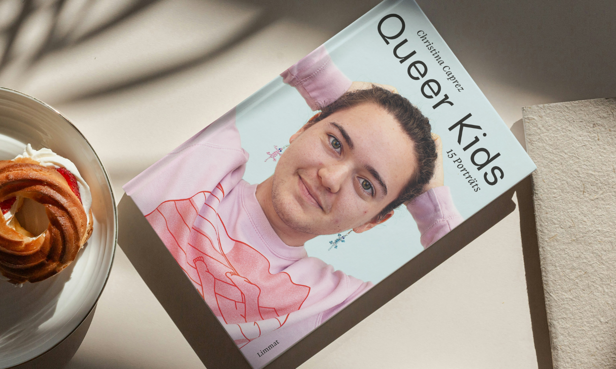 Das Buch Queer Kids liegt auf einem Tisch neben einem Teilchen. Auf dem Buchcover sehen wir eine Person, die direkt und freundlich in die Kamera schaut.