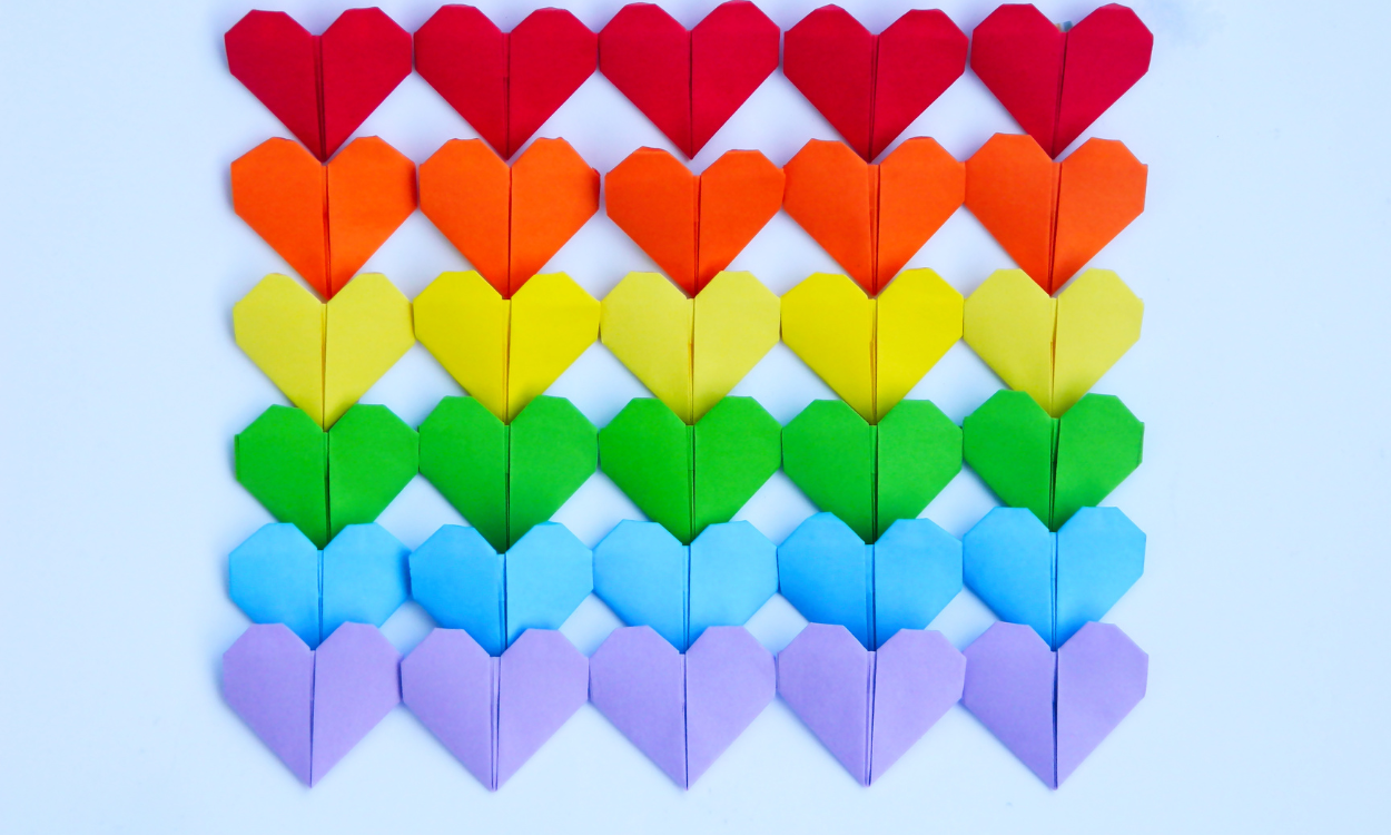 Kleine Papierherzen in Regenbogenfarben liegen auf einem hellblauen Hintergrund