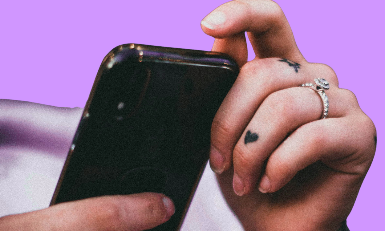 Eine Hand mit Tättowierungen und Ringen an den Fingern hält ein Handy vor einem lila Hintergrund.