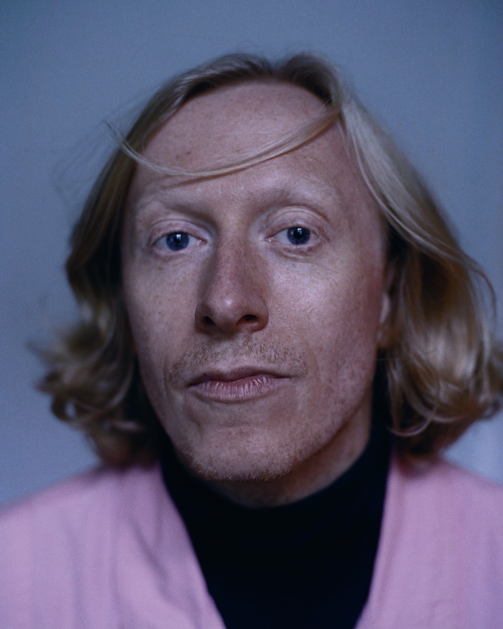 Der Autor mit den blonden, fast schulterlangem Haar schaut direkt in die Kamera. Er trägt ein rosa-schwarzes Oberteil