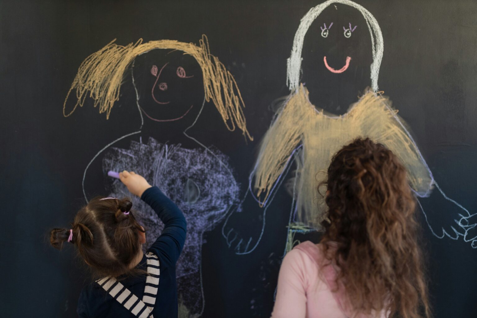 Zwei Kinder stehen vor einer Tafel und malen mit Kreide sich selbst mit bunter Kleidung und lächelnden Gesichtern